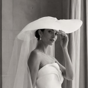 May include: A white wide-brimmed hat paired with a sheer, floor-length veil. The hat features a structured, rounded crown and a broad, sweeping brim. The veil is made of lightweight, translucent fabric that drapes elegantly from the hat's edge.