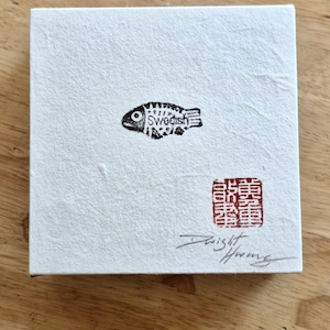 May include: A square piece of textured white paper featuring a small black ink illustration of a fish with the word Swedish printed on its body. A red square stamp with Asian characters and a handwritten signature in black ink are visible below.