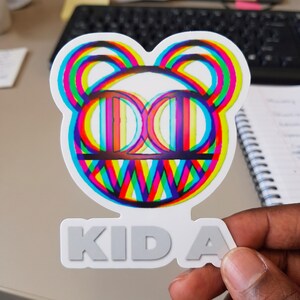 May include: A die-cut sticker featuring a stylized bear head graphic with a vibrant, multi-colored glitch effect in shades of cyan, magenta, and yellow. Below the bear head, the text KID A is printed in a bold, light gray sans-serif font.
