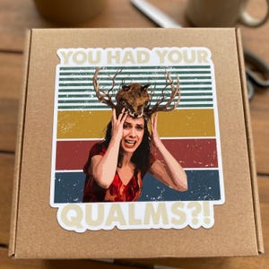 May include: A square sticker on a brown cardboard box featuring a person with a fox head and antlers on their head. The text reads YOU HAD YOUR QUALMS?! against a background of horizontal stripes in green, yellow, red, and blue with a distressed finish.