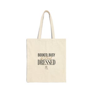 May include: Cream-colored tote bag with black text that reads "BOOKED, BUSY & BEST DRESSED". The bag has two handles and a simple design. A small illustration of a dress is below the word "DRESSED".