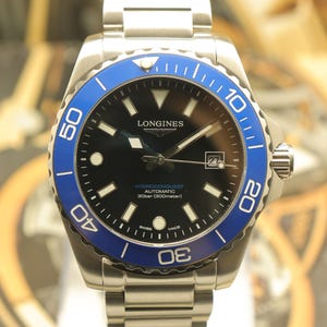 May include: A stainless steel Longines HydroConquest automatic watch with a black dial, blue rotating bezel, and a date window at the three o'clock position. The watch features luminous indices, silver hands, and a metal link bracelet strap.