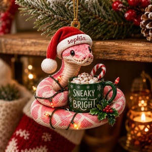 May include: A pink and white snake ornament wearing a red Santa hat with the name Sophia printed on the brim. The snake is coiled around a green mug filled with marshmallows and a candy cane, featuring the text Sneaky and Bright in white.