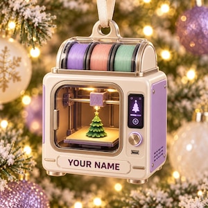 May include: A cream and purple 3D printer ornament featuring three filament spools in purple, pink, and teal on top. The front display shows a Christmas tree icon and the text YOUR NAME. A miniature green Christmas tree is inside the print chamber.