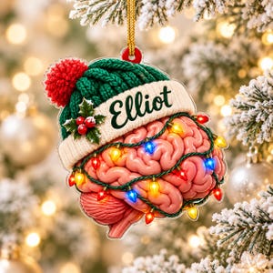 May include: A Christmas ornament shaped like a human brain wearing a green knit hat with a red pom-pom and a white brim featuring the name Elliot in black script. The brain is wrapped in a string of colorful glowing holiday lights.