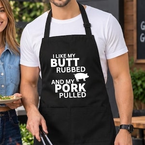 May include: A black apron featuring the white text I LIKE MY BUTT RUBBED AND MY PORK PULLED with a small pig graphic. The apron has a neck strap and a front pocket, designed for cooking and grilling tasks in a kitchen or outdoor barbecue setting.