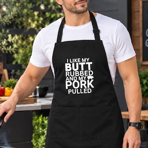 Może przedstawiać: Czarny fartuch z białym napisem I LIKE MY BUTT RUBBED AND MY PORK PULLED oraz małą ikoną świni. Fartuch posiada pasek na szyję, wiązania w talii i dwie przednie kieszenie, założony na gładki biały t-shirt podczas grillowania na świeżym powietrzu.