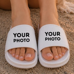 Peut inclure: Une paire de sandales à enfiler blanches dotées d'une large bride sur le dessus. Le texte "YOUR PHOTO" est imprimé en lettres capitales noires et grasses au centre de chaque bride. Les sandales ont une semelle profilée et une finition blanche.