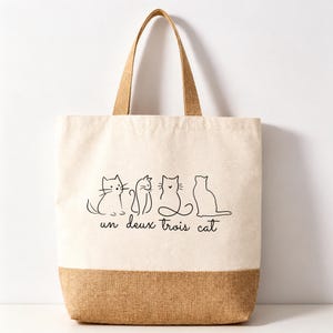 May include: A beige canvas tote bag with two brown handles and a brown base. The front features a black line art illustration of four cats in a row, with the text "un deux trois cat" printed in a cursive black font underneath the cat figures.
