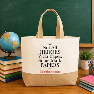 May include: A cream and tan canvas tote bag featuring the black printed text 'Not All HEROES Wear Capes, Some Mark PAPERS' with a small crown icon above the text and the placeholder 'Teacher name' printed in coral pink below the main quote.