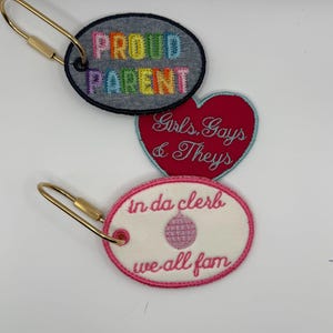 Pode incluir: Três chaveiros bordados com fechos dourados. Um é oval com "PROUD PARENT" em letras de arco-íris sobre um fundo de ganga. Outro é um coração vermelho com "Girls, Gays & Theys" em itálico. O último é oval com "in da clerb we all fam".