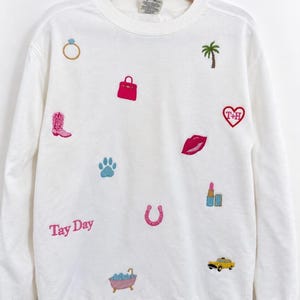 May include: A white crewneck sweatshirt featuring various embroidered patches including a ring, palm tree, pink handbag, cowboy boot, paw print, red heart with T+H, lips, lipstick, horseshoe, bathtub, yellow taxi, and the pink text Tay Day.
