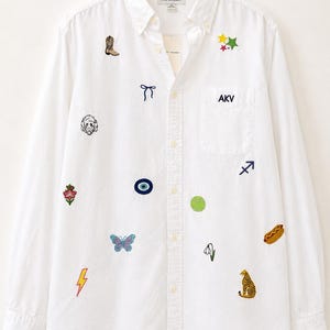May include: A white long-sleeve button-down shirt featuring various embroidered patches including a cowboy boot, a bow, a dog face, a flower, a butterfly, a lightning bolt, stars, the letters AKV, an eye, a tennis ball, a flower, a hot dog, and a leopard.