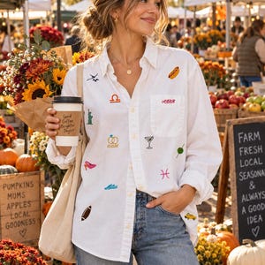 May include: A white button-down shirt featuring various small embroidered icons including a hot dog, a martini, a football, a taxi, a Libra symbol, a Pisces symbol, wedding rings with the date 10.10.26, a Statue of Liberty, and a pink high heel.