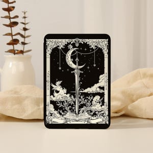 May include: A black rectangular e-reader case featuring a white line art illustration of a sword piercing an open book, set against a starry night sky with a crescent moon, clouds, and ornate decorative borders surrounding the central design.
