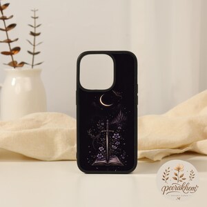 May include: A black smartphone case featuring a graphic illustration of an open book with a sword rising from its pages, surrounded by purple flowers, a deer silhouette, a crescent moon, and a distant castle under a starry night sky.