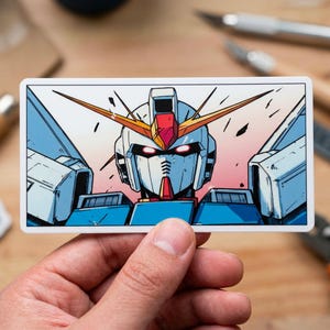 May include: A rectangular sticker featuring a detailed illustration of a Gundam robot's head. The robot has a white face, blue and red accents, and yellow antennae. The background transitions from pink to blue.