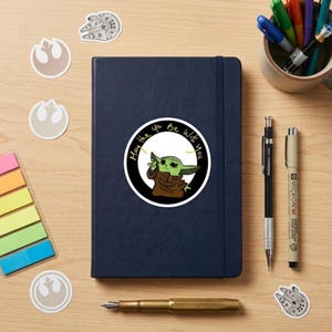 May include: A navy blue notebook with a round sticker of Grogu from The Mandalorian, with the text "May the 4th Be With You." The image includes Star Wars-themed stickers, pens, and a pen holder on a wooden surface.