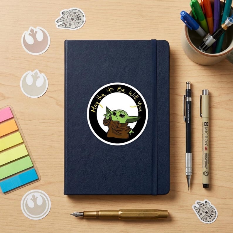 May include: A navy blue notebook with a round sticker of Grogu from The Mandalorian, with the text "May the 4th Be With You." The image includes Star Wars-themed stickers, pens, and a pen holder on a wooden surface.