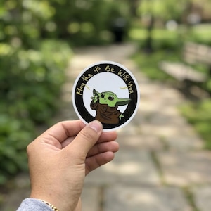 May include: A circular sticker with a green alien character in a brown robe, and the text "May the 4th Be With You" in a circular design. The character is making a hand gesture. The background is blurred.