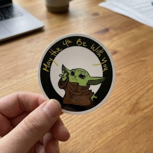May include: A circular sticker with a green cartoon character in a brown robe. The text "May the 4th Be With You" is written in gold around the character, set against a black and white background. The character is making a hand gesture.