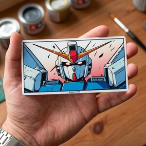 May include: A rectangular sticker featuring a stylized illustration of a Gundam robot's head. The robot has a white face, blue and red accents, and yellow antennae. The background transitions from pink to blue. The sticker has the text "BANDA / STKR".