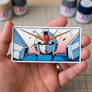 May include: A rectangular sticker featuring a detailed illustration of a Gundam robot's head and shoulders. The robot has a white, blue, red, and yellow color scheme, with a pink and orange gradient background. The sticker is held in a hand.