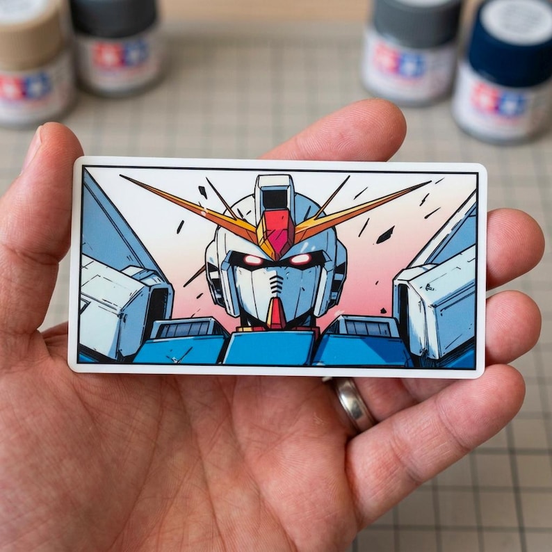 May include: A rectangular sticker featuring a detailed illustration of a Gundam robot's head and shoulders. The robot has a white, blue, red, and yellow color scheme, with a pink and orange gradient background. The sticker is held in a hand.