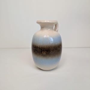 Może przedstawiać: Ceramiczny dzbanek o zaokrąglonym korpusie i małym uchwycie. Szkliwo charakteryzuje się przejściem kolorystycznym od kremowej bieli na górze i dole do szerokiego, ciemnobrązowego pasa pośrodku, podkreślonego delikatnymi jasnoniebieskimi paskami.