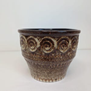 Op de afbeelding: Een bruine keramische plantenpot met een taps toelopende vorm en een brede rand. De buitenkant heeft een textuur met een herhalend patroon van reliëfspiralen rond het bovenste gedeelte en verticale ribbels langs de onderkant in aardetinten.
