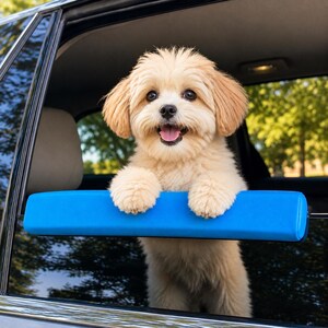 May include: A small, fluffy, light-tan puppy with floppy ears and a happy expression, resting its front paws on a bright blue, rectangular, padded car window sill protector. The puppy is positioned inside a vehicle, looking out the open window.