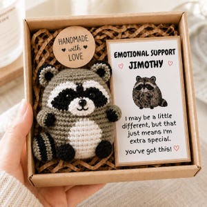 May include: A crochet raccoon plush toy with grey, white, and black yarn, nestled in a cardboard gift box with brown paper filler. A round tag reads Handmade with Love. A rectangular card features a raccoon illustration and the text Emotional Support Jimothy, I may be a little different, but that just means I'm extra special. you've got this!