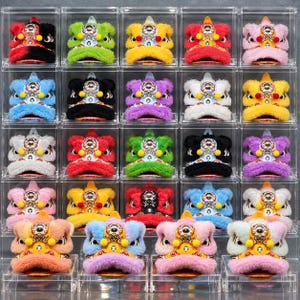 May include: A collection of twenty miniature lion dance masks displayed in individual clear acrylic boxes. Each mask features plush faux fur in various colors, a central metallic ornament, and yellow pom-poms, arranged in a grid pattern.