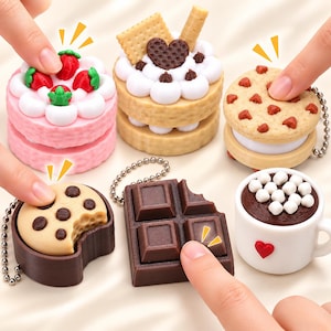 May include: A collection of miniature food-themed keychains including a strawberry cake, a heart-topped cake, a chocolate chip cookie, a bitten cookie, a chocolate bar, and a mug of hot cocoa with marshmallows, all featuring small metal chains.