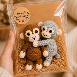 May include: Two small crocheted monkey dolls, one brown and one gray, with cream-colored faces, ears, hands, and feet. The monkeys are hugging and packaged in a clear plastic bag with a brown card backing featuring a sticker that says Handmade with love.