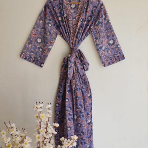 May include: A long-sleeved robe in a periwinkle blue hue featuring a repeating floral pattern in shades of white, dark blue, and muted orange. The garment includes a matching fabric tie belt at the waist and a wide collar with a contrasting border.