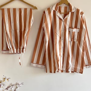May include: A brown and white striped pajama set. The long-sleeved shirt has a chest pocket and buttons. The trousers have a drawstring waist. The set is displayed on a wooden hanger. The measurements are in centimetres.