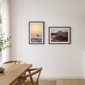 Könnte beinhalten: Zwei gerahmte Landschaftsfotografien hängen an einer weißen Wand. Der linke Rahmen zeigt ein Boot auf dem Meer bei Sonnenuntergang. Der rechte Rahmen zeigt eine Bergkette mit bewölktem Himmel. Die Rahmen sind schwarz.