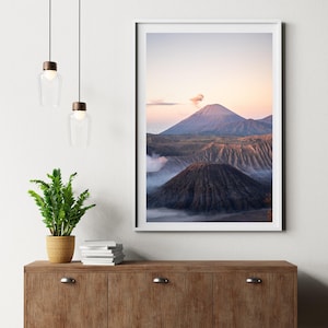 Può includere: Una stampa incorniciata di un paesaggio montano all'alba. L'immagine mostra un grande vulcano marrone scuro in primo piano, con altre montagne e nuvole sullo sfondo. Il cielo passa dal rosa al blu.