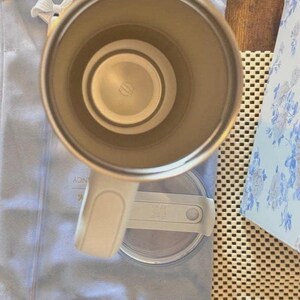 Puede incluir: Una vista desde arriba de un vaso t&eacute;rmico de color claro con interior met&aacute;lico y asa. El vaso est&aacute; colocado sobre su tapa circular transparente, que cuenta con un mecanismo de cierre deslizante. La base interior muestra un logotipo circular.