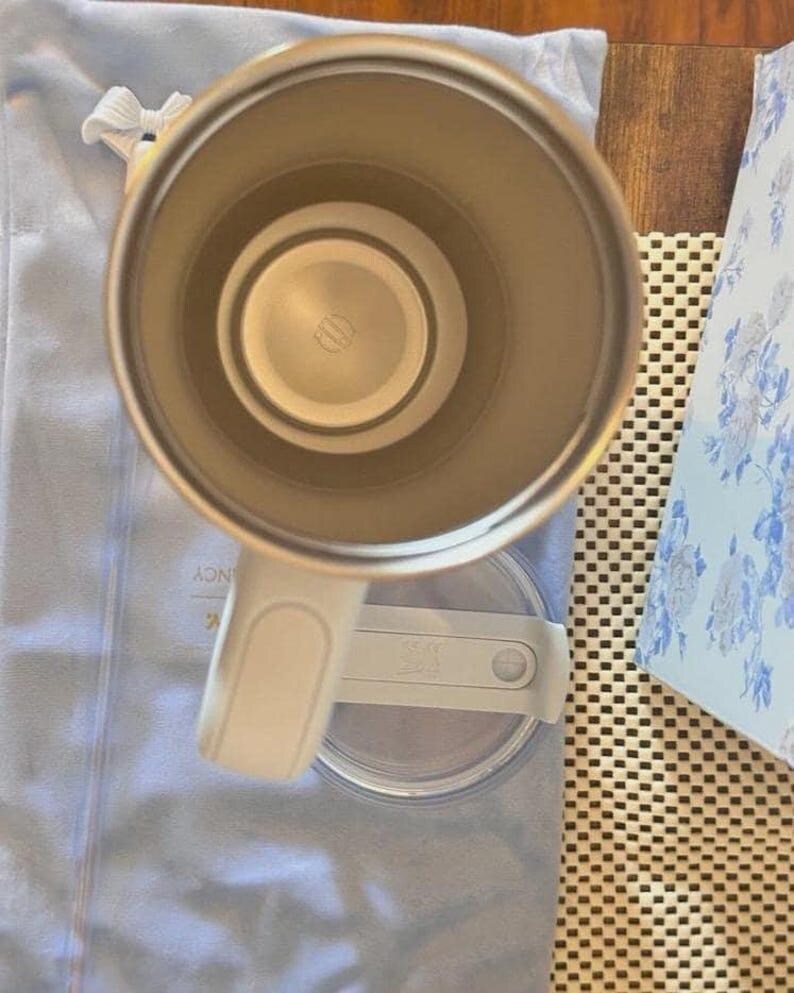 Puede incluir: Una vista desde arriba de un vaso t&eacute;rmico de color claro con interior met&aacute;lico y asa. El vaso est&aacute; colocado sobre su tapa circular transparente, que cuenta con un mecanismo de cierre deslizante. La base interior muestra un logotipo circular.