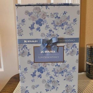 Puede incluir: Una caja de producto rectangular con un estampado floral en azul claro y blanco. Una cinta azul horizontal con un lazo central rodea el centro, mostrando el texto STANLEY y LOVESHACKFANCY. Una etiqueta rectangular tambi&eacute;n dice STANLEY LOVESHACKFANCY.