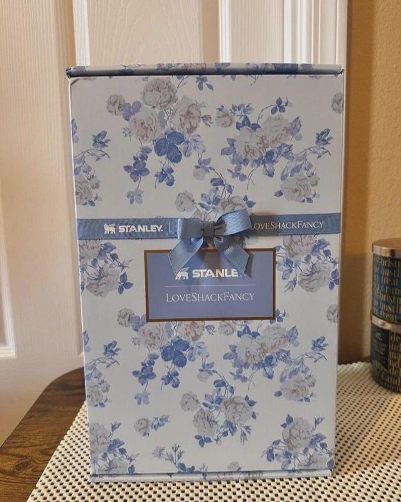 Puede incluir: Una caja de producto rectangular con un estampado floral en azul claro y blanco. Una cinta azul horizontal con un lazo central rodea el centro, mostrando el texto STANLEY y LOVESHACKFANCY. Una etiqueta rectangular tambi&eacute;n dice STANLEY LOVESHACKFANCY.