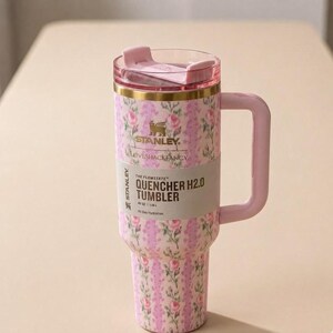 May include: A pink floral Stanley Quencher H2.0 tumbler with a handle. The tumbler has a pink lid and handle, and a floral pattern in pink and white. The text "LOVESHACKFANCY" and "QUENCHER H2.0 TUMBLER" are visible.