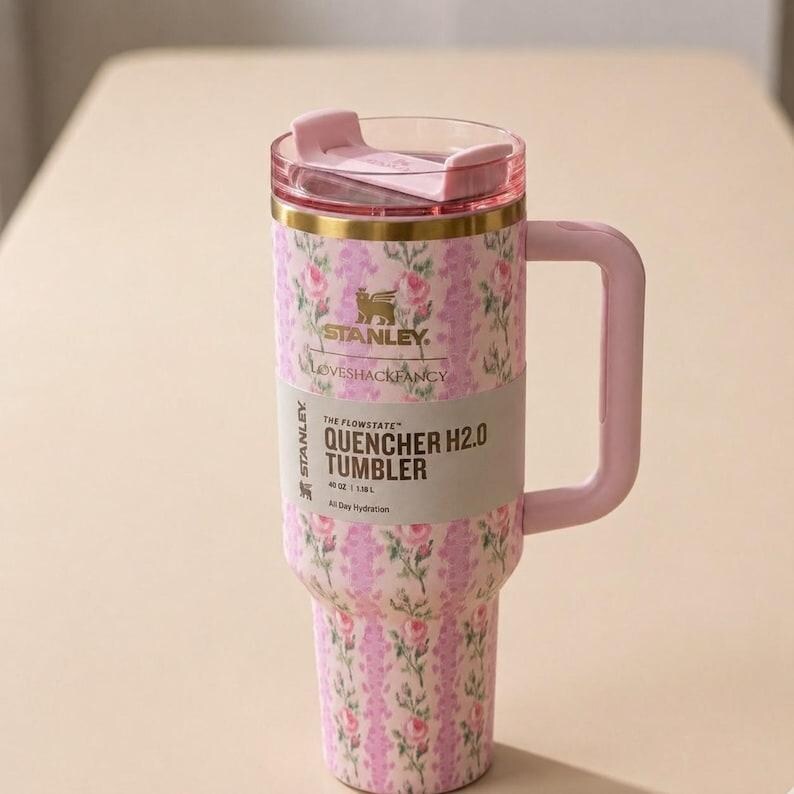 May include: A pink floral Stanley Quencher H2.0 tumbler with a handle. The tumbler has a pink lid and handle, and a floral pattern in pink and white. The text "LOVESHACKFANCY" and "QUENCHER H2.0 TUMBLER" are visible.