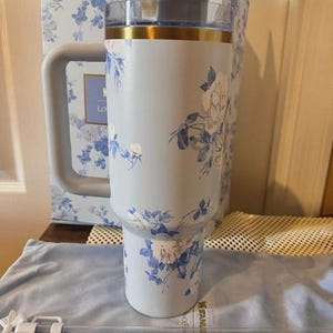 Puede incluir: Un vaso t&eacute;rmico de color azul claro con un estampado floral de rosas blancas y azules. El vaso tiene un asa lateral grande, una tapa transparente con un borde dorado y una pajita transparente con una flor blanca. La marca Stanley es visible.