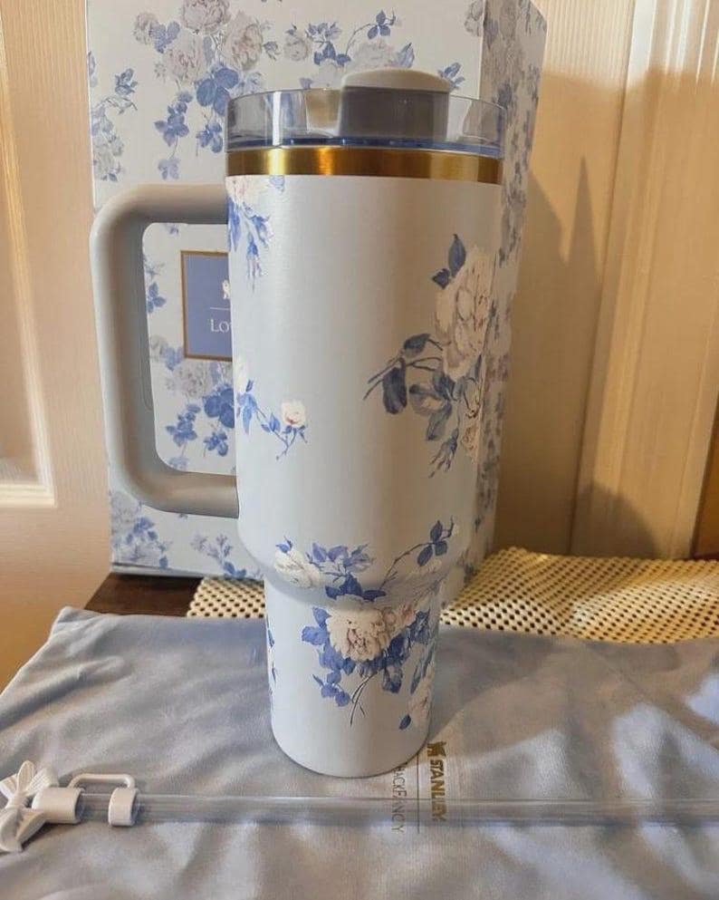 Puede incluir: Un vaso t&eacute;rmico de color azul claro con un estampado floral de rosas blancas y azules. El vaso tiene un asa lateral grande, una tapa transparente con un borde dorado y una pajita transparente con una flor blanca. La marca Stanley es visible.