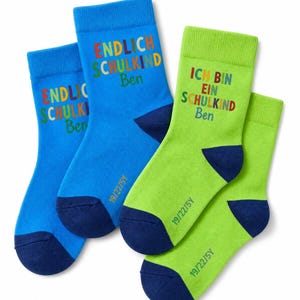 Könnte beinhalten: Zwei Paar Socken in Blau und Limettengrün mit dunkelblauen Fersen und Zehen. Die blauen Socken tragen den Text ENDLICH SCHULKIND Ben in bunten Buchstaben. Die limettengrünen Socken tragen den Text ICH BIN EIN SCHULKIND Ben. Die Größe 19/22/5Y ist aufgedruckt.