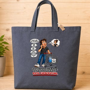 Könnte beinhalten: Eine dunkelgraue Stofftasche mit einer Cartoon-Illustration einer Person mit Einkaufswagen beim Telefonieren. Der Text lautet: "SCHATZ, WAS SOLL ICH NEHMEN? ODER DAS? UND DAS? ODER DOCH DAS? WAS MEINST DU? 2 STUNDEN EINKAUFEN. 2 SACHEN GEKAUFT. 100% EHEBERATUNG."