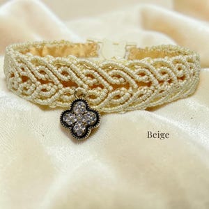 Puede incluir: Collar de macramé beige con un dije de trébol de cuatro hojas. El collar presenta un diseño tejido con una base crema y un interior dorado. El dije es plateado con piedras claras y brillantes. La palabra "Beige" está impresa en la parte inferior derecha.